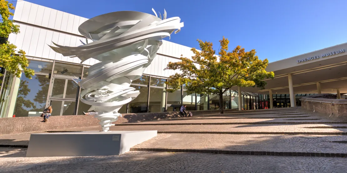 Skultptur in Form eines Wirbels vor dem Eingang des Sprengel Museums, im Hintergrund herbstlich belaubter Baum.
